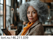 An elegant woman with grey, thick and voluminous curls, sitting in a cozy room with warm natural light. She holds a tablet in her hands and looks with confidence and dignity. Her image is emphasized by a stylish coat and a warm knitted scarf of mustard color. The background is slightly blurred, creating an atmosphere of coziness and concentration, perhaps in a cafe, library or workshop. Стоковое AI-изображение, AI-художник Анна Кривицкая / Фотобанк Лори
