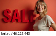 A cheerful woman stands confidently beside a large red sale sign against a vibrant orange background, promoting a special offer with enthusiasm. Seasonal discounts and promos. Стоковое AI-изображение, AI-художник Sergey Grishin / Фотобанк Лори