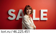 A cheerful woman poses with her arms crossed against a bold red backdrop. The large white letters spell out \'SALE,\' emphasizing a festive shopping environment. Seasonal discounts and promos. Стоковое AI-изображение, AI-художник Sergey Grishin / Фотобанк Лори