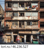 An old apartment building in an urban setting. The building's façade looks dilapidated: the walls show signs of time - peeling paint, cracks, dirt and rust. The balconies are partially covered with metal bars, and colorful laundry is hung on them.On the ground floors there are small shops and stalls with various goods, among which carpets, food and household items are visible. Open doors and windows create a sense of everyday life, despite the general chaotic appearance of the building. Wires run haphazardly along the walls and between buildings, emphasizing the atmosphere of a densely populated and not very well-kept area.This scene conveys the authentic color of city life, showing a combination of everyday life, trade and characteristic architectural details of old residential buildings. Стоковое AI-изображение, AI-художник Анна Кривицкая / Фотобанк Лори