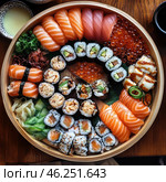 A beautifully presented platter of sushi and rolls on a round wooden plate. In the center are various maki rolls with avocado, salmon, tuna, eel and other ingredients. Along the edges are neatly laid out nigiri with salmon and tuna, as well as pieces of grilled eel with sauce. Red caviar is visible in the corner, adding a bright accent. In addition to the main dish, there is a garnish: wasabi, pickled ginger, green onions and seaweed. Nearby are cups with soy sauce and, perhaps, sake or green tea. The composition looks aesthetically pleasing and appetizing. Стоковое AI-изображение, AI-художник Анна Кривицкая / Фотобанк Лори