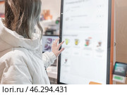 A woman in a white jacket chooses drinks and orders food for dessert in a restaurant or pizzeria coffee shop through a touchscreen screen on her own. The restaurant menu is displayed on the screen in the terminal. Customers frequently using selfservice kiosks in modern retail environments is notable. Стоковое фото, фотограф Владимир Разгуляев / Фотобанк Лори