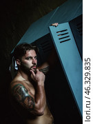 A muscular man stands in a dark, indoor locker room, contemplating while leaning against a blue locker. His thoughtful expression suggests introspection amidst raw strength and artistry. Стоковое фото, фотограф Aleksandr Golubev / Фотобанк Лори