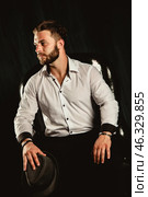 A man with a well-groomed beard sits confidently in a black chair, wearing a crisp white shirt and holding a hat. The moody lighting highlights his thoughtful expression and fashionable style. Стоковое фото, фотограф Aleksandr Golubev / Фотобанк Лори