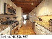 A cozy country-style kitchen with modern touches. The narrow but functional space is decorated in warm colors, with a wooden ceiling with exposed beams and natural wood floors. White kitchen cabinets with classic handles harmonize with granite countertops, and a backsplash made of light tiles.On the left are modern stainless steel kitchen appliances, including a gas stove with oven and microwave. On the right, on the countertop, there is a knife block and kitchen utensils. At the end of the kitchen, you can see the living area with large windows that let in natural light, as well as a houseplant, creating a cozy atmosphere. Стоковое AI-изображение, AI-художник Анна Кривицкая / Фотобанк Лори