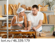 In bakery, male visitor and female salesperson work with paper documents and count using phone. Стоковое фото, фотограф Яков Филимонов / Фотобанк Лори