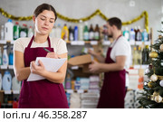 Attentive saleswoman restock of goods, checking papers, male assistant laying out goods in background in small household store decorated for New Year celebration. Стоковое фото, фотограф Яков Филимонов / Фотобанк Лори
