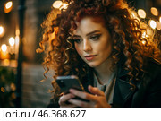 A businesswoman with curly red hair and freckles. She's wearing a coat. In the hands of a smartphone. A small business entrepreneur looks at her mobile phone. Стоковое AI-изображение, AI-художник Photojuli / Фотобанк Лори