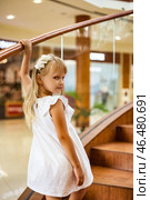 In a vibrant shopping center, a charming girl with long blonde hair ascends a wooden staircase. She wears a white dress and a delicate flower hair accessory, exuding playfulness and curiosity. Стоковое фото, фотограф Aleksandr Golubev / Фотобанк Лори