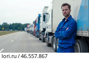 Truck driver standing confidently beside truck on highway with long line of parked trucks in background, wearing blue work uniform, looking serious, transportation industry, logistics, cargo delivery. Стоковое AI-изображение, AI-художник Павел Муравьев / Фотобанк Лори