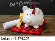 Копилка в форме выпускного берета с дипломом и калькулятором на коричневом деревянном столе перед меловой доской. Концепция студенческого займа. Стоковое фото, фотограф Pixel_shot / Фотобанк Лори
