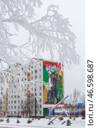 Город Тында, Амурская область, Россия. Вид на большое граффити на стене жилого дома, посвящённое строителям Байкало-Амурской магистрали (БАМ) и города Тынды. Мурал с изображением молодых энергичных людей, идущих на стройку. На заснеженной улице скульптурная композиция в виде серпа и молота. На переднем плане, покрытые инеем, ветви дерева. (2024 год). Редакционное фото, фотограф Андрей Степанов / Фотобанк Лори