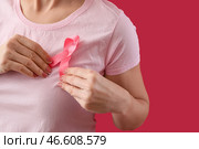 Женщина поправляет розовую ленту осознания на груди на красном фоне, крупным планом. Концепция осведомленности о раке молочной железы. Стоковое фото, фотограф Pixel_shot / Фотобанк Лори