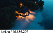 Romantic beachfront cottage illuminated at night surrounded by calm waters and palm trees in a tropical paradise. Стоковое AI-изображение, AI-художник Михаил Коханчиков / Фотобанк Лори