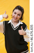 A young girl with short dark hair braided on top of her head. She smiles and makes a thumbs up gesture, expressing positive emotions. The girl is wearing a white shirt with a black knitted vest with a contrasting white trim. In the background is a bright yellow wall, creating a cheerful and energetic atmosphere. Стоковое AI-изображение, AI-художник Анна Кривицкая / Фотобанк Лори