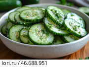 Fresh cucumber salad, thinly sliced ​​and dressed with herbs, sesame seeds and a light dressing. The dish is served in a deep white plate with a speckled pattern, standing on a wooden surface. The freshness and juiciness of the cucumbers are emphasized by the shiny surface of the slices, and the green herbs add flavor and color. In the background, whole cucumbers and herbs are blurred, creating an effect of naturalness and freshness. Стоковое AI-изображение, AI-художник Анна Кривицкая / Фотобанк Лори