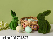 Пасхальные кролики и плетеная корзина с крашеными яйцами на столе на зеленом фоне. Стоковое фото, фотограф Pixel_shot / Фотобанк Лори
