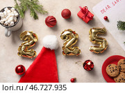 Цифра 2025 из фольгированных шаров с шапкой Санты, рождественскими шарами, сладкими печеньями, подарочной коробкой и чашкой какао на бежевом фоне. Стоковое фото, фотограф Pixel_shot / Фотобанк Лори