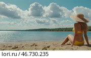 A woman on vacation, sitting in a swimsuit and a wide-brimmed straw hat on the beach on a sunny day and looking at the sea. Стоковое AI-изображение, AI-художник Photojuli / Фотобанк Лори