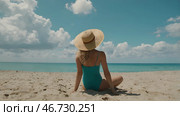 A woman on vacation, sitting in a swimsuit and a wide-brimmed straw hat on the beach on a sunny day and looking at the sea. Стоковое AI-изображение, AI-художник Photojuli / Фотобанк Лори
