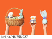 Женские руки держат корзину с яйцами, пасхальным куличом и подарком на оранжевом фоне. Стоковое фото, фотограф Pixel_shot / Фотобанк Лори