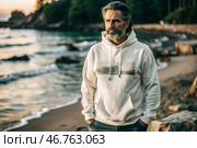 A handsome middle-aged man with a beard wearing a white hoodie on the beach. Template layout for hoodie design, print area layout for logo or design. Стоковое AI-изображение, AI-художник Photojuli / Фотобанк Лори