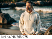 A handsome middle-aged man with a beard wearing a white hoodie on the beach. Template layout for hoodie design, print area layout for logo or design. Стоковое AI-изображение, AI-художник Photojuli / Фотобанк Лори