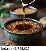 A close-up of a beautiful ceramic bowl filled with granulated brown sugar or caramel granules, into which a thick dark liquid reminiscent of honey, syrup or molasses is poured. The background is slightly blurred, creating a cozy and warm atmosphere. A second bowl and wooden elements in the background complete the composition, creating a sense of traditional cuisine and natural products. Стоковое AI-изображение, AI-художник Анна Кривицкая / Фотобанк Лори