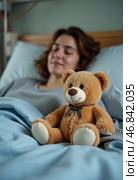 Brightened by a cherished teddy bear, a resilient woman lies in bed at the hospital, finding comfort amid her struggle. Hope and care surround her during this challenging time. Стоковое AI-изображение, AI-художник Юлия Петровская / Фотобанк Лори