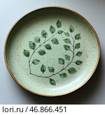 Ceramic plate with an elegant natural pattern. It has a light green base with dark specks, creating the effect of natural glaze. In the center of the plate there is a branch with green leaves, made in a watercolor or botanical style. The edges of the plate are slightly aged, which gives it a rustic, vintage look. The background is minimalist, which emphasizes the sophistication and elegance of the item. Стоковое AI-изображение, AI-художник Анна Кривицкая / Фотобанк Лори