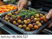 A close-up of a black plastic container with multi-colored olives and black olives, garnished with sprigs of fresh rosemary. The olives look fresh, shiny, soaked in oil or marinade. In the distance, you can see a plate of bright cherry tomatoes, creating an additional color accent. Someone's hands are carefully holding the container, perhaps serving it or choosing it from among other products. The image conveys an atmosphere of gastronomic delights, Mediterranean cuisine and a love of natural products. Стоковое AI-изображение, AI-художник Анна Кривицкая / Фотобанк Лори