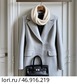 Elegant grey coat with cream scarf and black handbag displayed against a minimalist wall backdrop. Стоковое AI-изображение, AI-художник Сергей Толмачёв / Фотобанк Лори