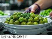 A close-up shot of green olives in a deep white ceramic bowl. They are coated in a layer of aromatic oil, giving them an appetizing shine. The olives are topped with fresh sprigs of thyme, which adds flavor and piquancy to the dish. The frame also shows a hand holding a spoon, which creates a sense of the process of preparing or serving the dish. The background is slightly blurred, with elements of the kitchen and additional plates with herbs, which enhances the gastronomic atmosphere. Стоковое AI-изображение, AI-художник Анна Кривицкая / Фотобанк Лори