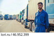 Truck driver in blue uniform standing near truck on highway with long row of trucks in traffic jam, serious look, transportation, freight delivery, logistics, cargo, trucking industry, supply chain. Стоковое AI-изображение, AI-художник Павел Муравьев / Фотобанк Лори