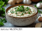 A bowl of thick, creamy sauce or dip, garnished with freshly chopped parsley and a sprinkle of black pepper. The dish has a natural, ceramic look with a speckled pattern, adding to the homey feel. Ingredients such as garlic, herbs and perhaps ginger root are arranged around the table, creating a natural, culinary atmosphere. The background is neutral, slightly washed out, which highlights the main dish. Стоковое AI-изображение, AI-художник Анна Кривицкая / Фотобанк Лори
