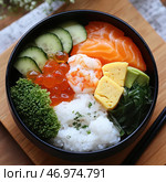 Traditional Japanese dish - kaisen-don (seafood on rice). Fresh ingredients are beautifully laid out in a black bowl: slices of juicy salmon, tender shrimp, bright salmon roe (ikura), sliced ​​cucumber, avocado, Japanese omelet (tamago), wakame seaweed, broccoli and white rice, garnished with finely chopped green onions. The dish looks balanced in color and composition, combining seafood, vegetables and grains. In the background you can see a bamboo board and chopsticks, creating the atmosphere of traditional Japanese cuisine. Стоковое AI-изображение, AI-художник Анна Кривицкая / Фотобанк Лори