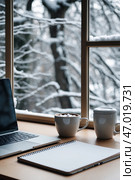 Cozy workspace by the window overlooking a snowy landscape during winter mornings. Стоковое AI-изображение, AI-художник Сергей Толмачёв / Фотобанк Лори