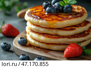 A delicious stack of fluffy golden pancakes drizzled with viscous honey or maple syrup. The top pancake is topped with fresh blueberries and mint leaves, while juicy strawberries surround it, adding even more brightness and freshness to the scene. The dish is placed on a wooden board, creating a cozy, homey atmosphere. The light falls softly on the dessert, emphasizing its texture and creating a warm gastronomic accent. Стоковое AI-изображение, AI-художник Анна Кривицкая / Фотобанк Лори