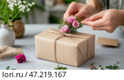 Wrapping a gift in biodegradable paper adorned with flowers in a cozy workspace. Упаковка подарка в биоразлагаемую бумагу, украшенную цветами, в уютном рабочем пространстве. Стоковое AI-изображение, AI-художник Katsiaryna Repich / Фотобанк Лори