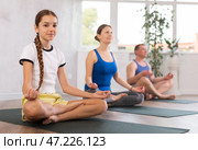 Middle-aged woman, man, preteen girl and boy practicing Padmasana with crossed legs on mats during family Hatha yoga class in wellness studio. Стоковое фото, фотограф Яков Филимонов / Фотобанк Лори