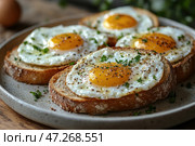 A delicious breakfast: fried eggs served on toasted slices of bread. The yolks are a rich golden color, while the whites frame them neatly, creating a beautiful contrast. The dish is garnished with fresh herbs, perhaps parsley, and seasoned with ground black pepper and salt. All this is placed on a stylish ceramic plate with a natural texture, emphasizing the homely atmosphere. In the background, blurred details of the kitchen are visible, creating a warm atmosphere. Стоковое AI-изображение, AI-художник Анна Кривицкая / Фотобанк Лори