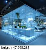 A futuristic glass room that can be a restaurant or an exhibition stand. The entire interior is designed in a minimalist style with a predominance of white and blue lighting, creating the effect of purity and modernity. The space is surrounded by glass walls, which makes it visually airy and transparent. Inside you can see tables, chairs, green plants, shelves with dishes and decorative elements. Pendant lamps add coziness, and living plants soften the strict high-tech design. Стоковое AI-изображение, AI-художник Анна Кривицкая / Фотобанк Лори