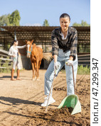 Smiling Asian girl worker scoops horse droppings into shovel and cleans up cattle. Стоковое фото, фотограф Яков Филимонов / Фотобанк Лори