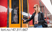 Young blond woman in jeans, shirt and leather jacket wearing bag and sunglass, presses door button of modern speed train to embark on train station platform. Travel and transportation. Стоковое фото, фотограф Matej Kastelic / Фотобанк Лори