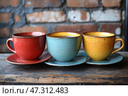 Three ceramic cups and saucers, standing on a wooden surface against a brick wall. Each cup has a rich color and vintage texture with small specks: red on the left, blue in the center and yellow on the right. The inner walls of the cups are covered with a glossy glaze, which gives additional warmth and coziness to the composition. This image is associated with soulful communication, a cozy coffee break or a warm start to the day. Стоковое AI-изображение, AI-художник Анна Кривицкая / Фотобанк Лори