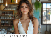 A young woman with natural beauty and a soft, friendly aura. Her warm gaze is directed straight at the camera, creating a sense of sincerity and a lively presence. Her hair is slightly tousled, giving the image lightness and freedom. A light tan, blush and minimal makeup emphasize her natural attractiveness. She is dressed in a white top with a deep neckline, which adds to the summer mood. The background is a cozy cafe or store, with a blurred interior and soft light. This is a portrait of a young modern heroine, living in harmony with herself and the world around her. Стоковое AI-изображение, AI-художник Анна Кривицкая / Фотобанк Лори