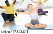 Girl conducts Pilates multinational group sitting with elastic band in hands above head body bends. Стоковое видео, видеограф Яков Филимонов / Фотобанк Лори