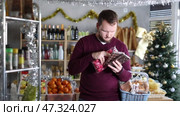 In New Year-themed grocery store, young man holding chocolate bars and bag of candies, considering adding to basket of holiday treats and snacks. Стоковое видео, видеограф Яков Филимонов / Фотобанк Лори