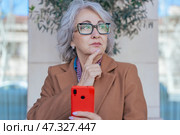 Thoughtful senior woman with smartphone looking away in an urban environment. Стоковое фото, фотограф Raquel Corcoles Mendez / Фотобанк Лори