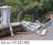 A collapsed section of a balustrade, made in the classical style with white balusters. It has partially collapsed down from a terrace or path framed by stonework. The debris lies on a paved path, part of the fence is still in place. It is clear that the area was fenced off with red and white tape for safety. There is green vegetation, trees and bushes around. The place looks like a park or embankment, perhaps this is Simeiz, Crimea. Стоковое фото, фотограф Анна Кривицкая / Фотобанк Лори